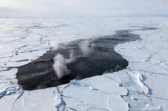 A wide channel of open dark water cut through endless white pack ice, steam rising into cold air
